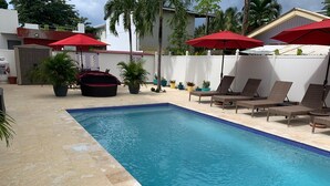 A swimming pool with lounge chairs and red umbrellas, surrounded by potted plants and a white fence.