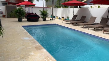 A swimming pool with lounge chairs and red umbrellas, surrounded by potted plants and a white fence.