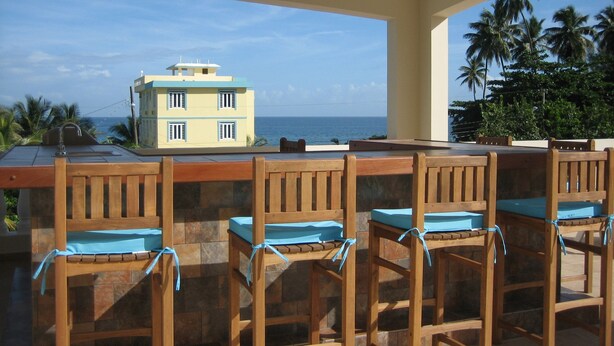A covered outdoor seating area with wooden chairs and a table, overlooking a building and the ocean.