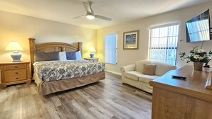 Un dormitorio con cama, mesitas de noche con lámparas, cabecero de madera, un sofá, un televisor y una ventana con persianas.