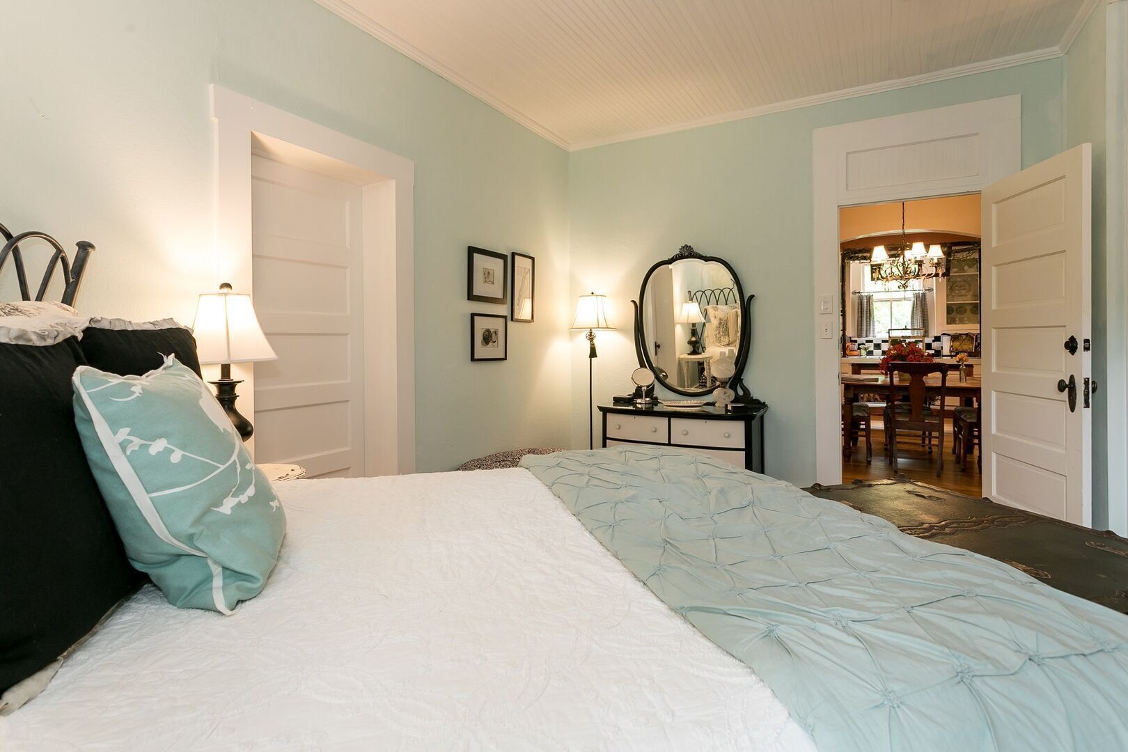 A bedroom with a bed, bedside lamps, a dresser, a mirror, and a view into the kitchen.