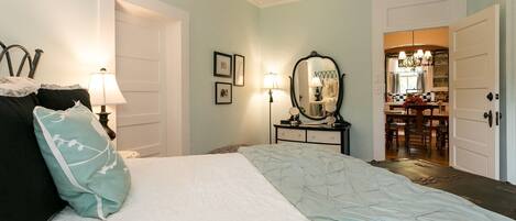A bedroom with a bed, bedside lamps, a dresser, a mirror, and a view into the kitchen.
