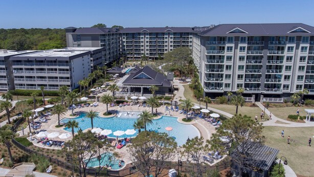 Um hotel com vários edifícios, uma piscina central rodeada por espreguiçadeiras e guarda-sóis, e uma área ajardinada bem cuidada.