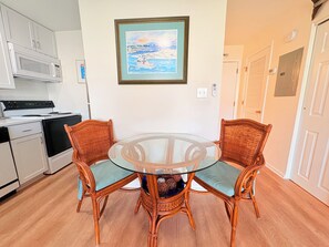 A kitchen with white cabinets, a microwave, and a stove. There is a round glass table with two wicker chairs. A framed painting hangs on the wall above the table.