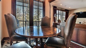 Una mesa de madera redonda con cuatro sillas tapizadas, ventanas amplias con persianas y una zona de bar con televisor.