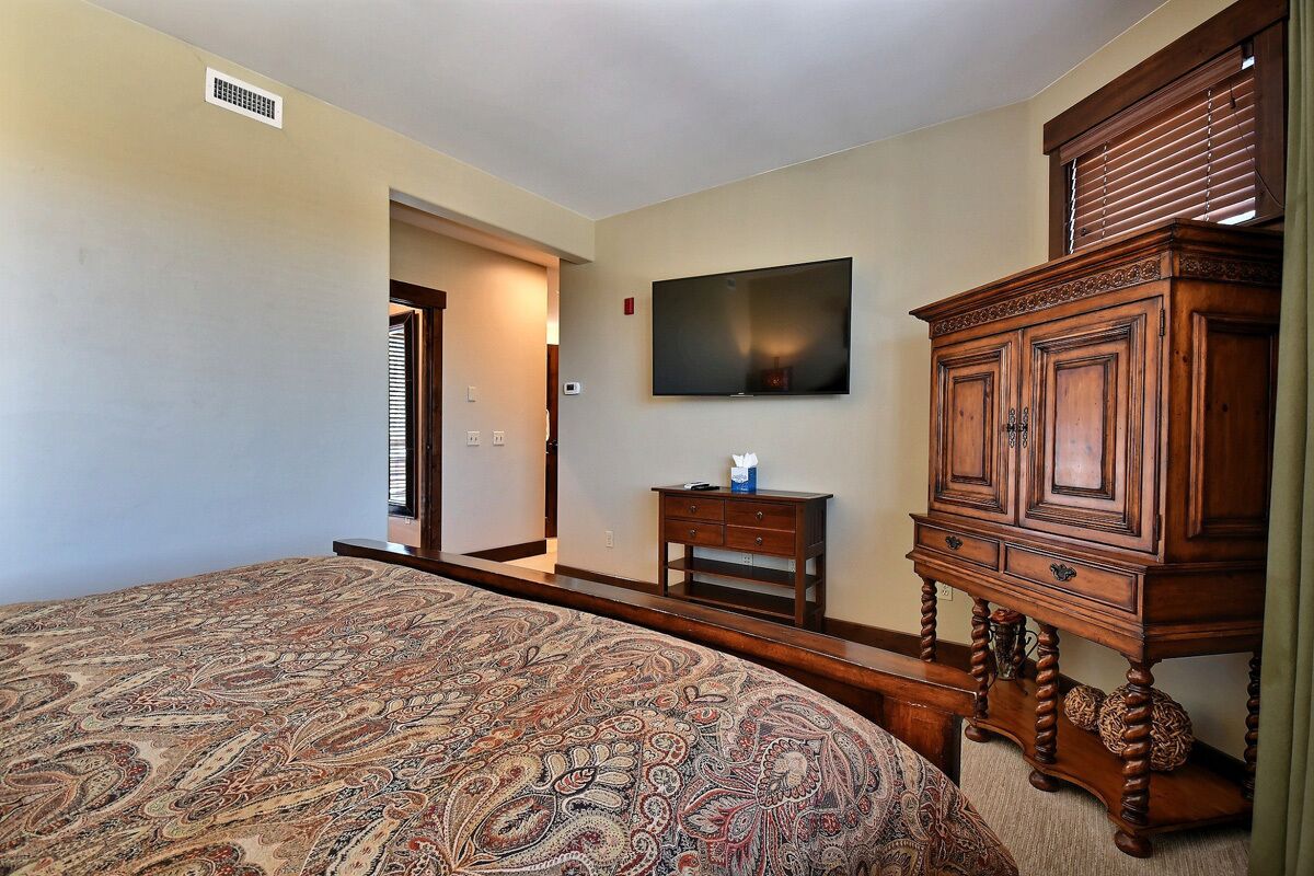 Een slaapkamer met een groot bed, een aan de muur bevestigde televisie, een houten kast en een deur naar een andere kamer.