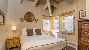 A bedroom with a bed, wooden headboard, a round mirror, a nightstand, a lamp, and a window with a view of trees. - Seasonal Pool | Pickleball | Hot Tub (Ketchum)
