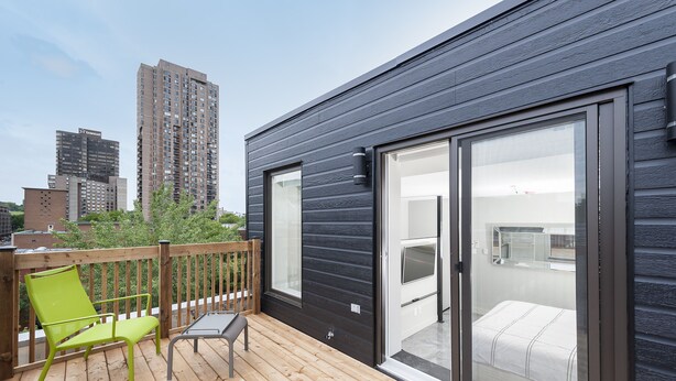 A modern building with a dark exterior, a balcony with a wooden deck, and a sliding glass door leading to a bedroom with a bed. - Duplex Loft with Private Terrace, Parc Avenue (Montréal)