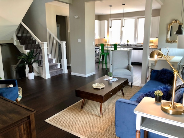 Un salon moderne avec un escalier, une cuisine en arrière-plan et un coin repas.