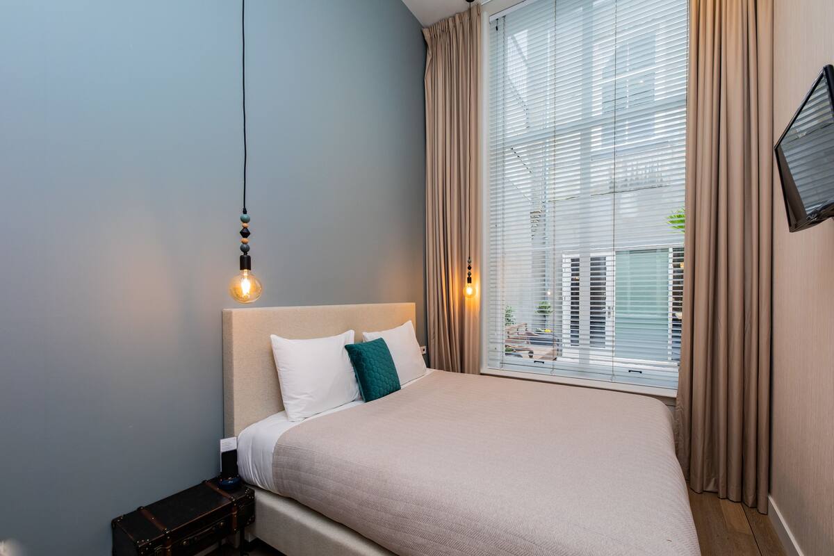 A neatly made bed with a headboard, a bedside table with a lamp, and a window with curtains.