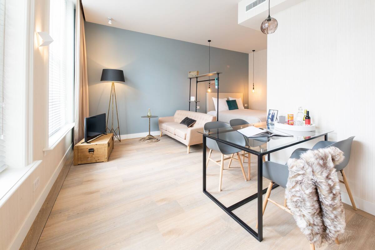 A modern, well-lit bedroom with a bed, bedside table, lamp, and a glass table with chairs.