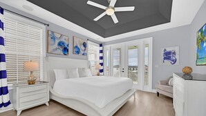 A bedroom with a large bed, a nightstand with a lamp, a dresser, and a ceiling fan. There are windows with blinds, a door leading to a balcony, and artwork on the walls.