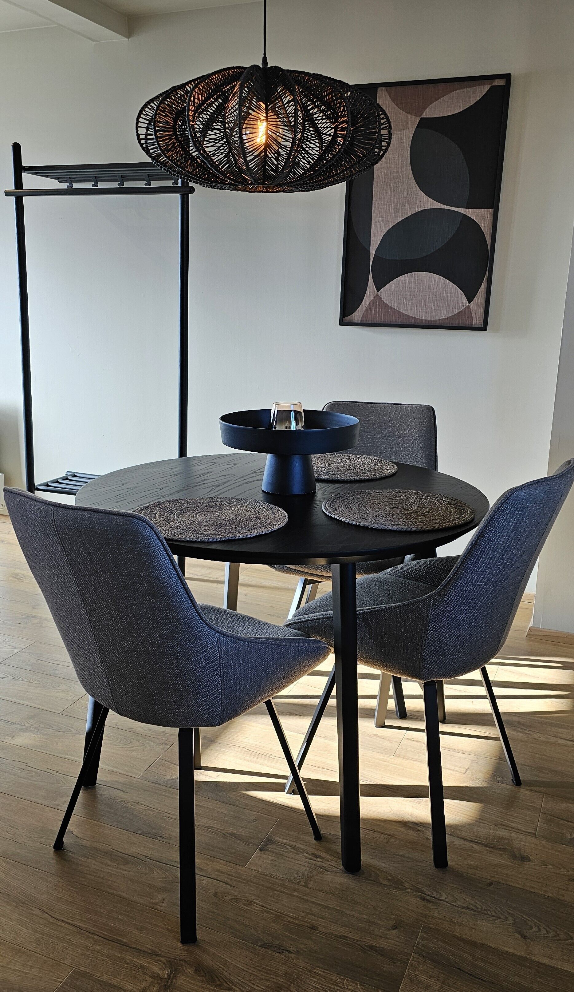 Un comedor con una mesa redonda, cuatro sillas, una lámpara colgante negra y una obra de arte en la pared.