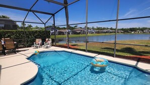 A swimming pool with a metal safety ladder, surrounded by a covered area with chairs and a view of a lake and houses in the background. - Brand new private pool with lake view minutes to the parks! (Kissimmee)