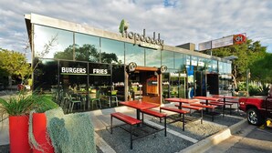 Un restaurante moderno con fachada de vidrio, mesas al aire libre y un cartel que anuncia hamburguesas y papas fritas.