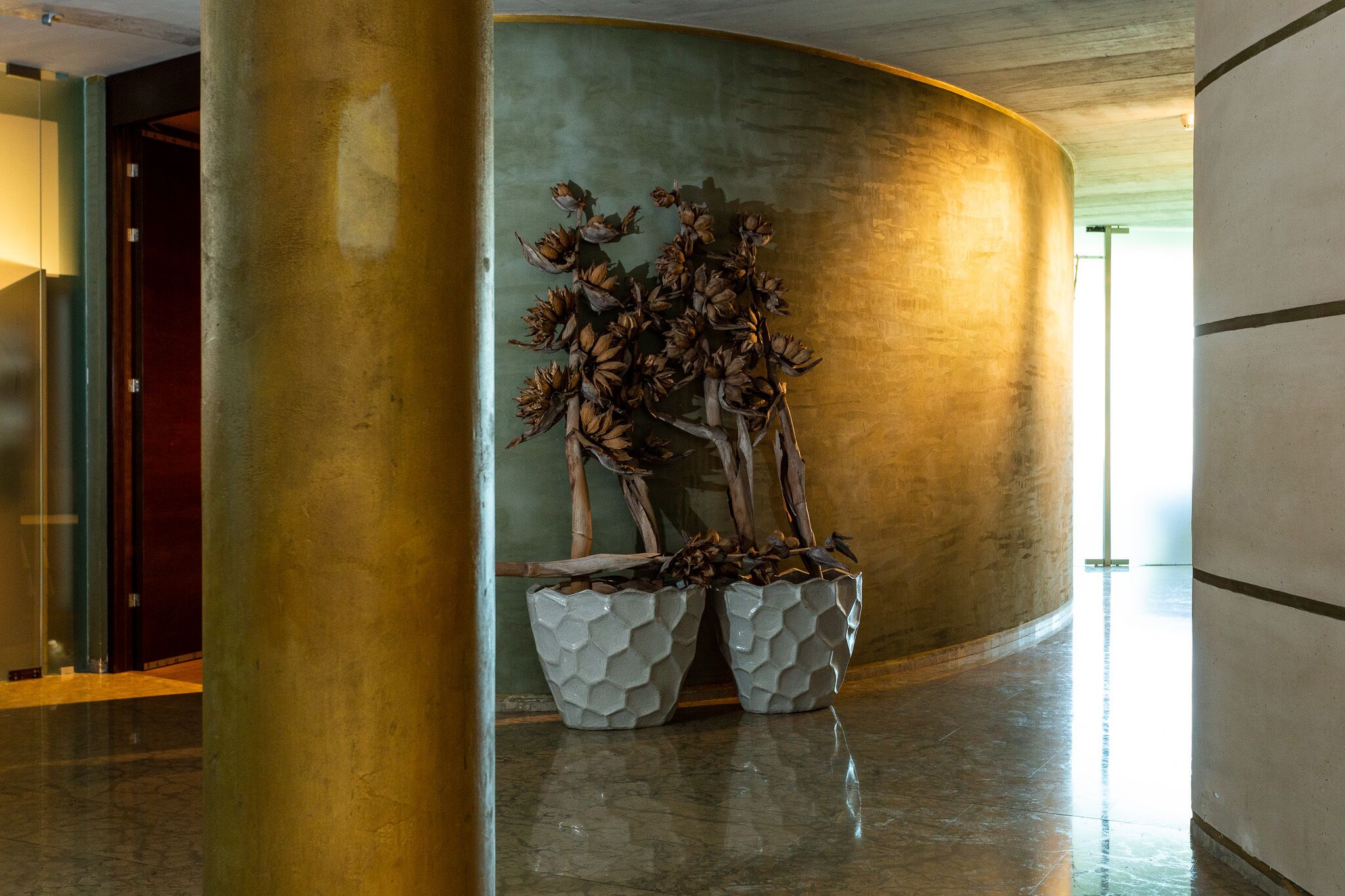 En moderne lobby med en buet væg, en stor søjle og to sekskantede plantekasser med tørrede planter.