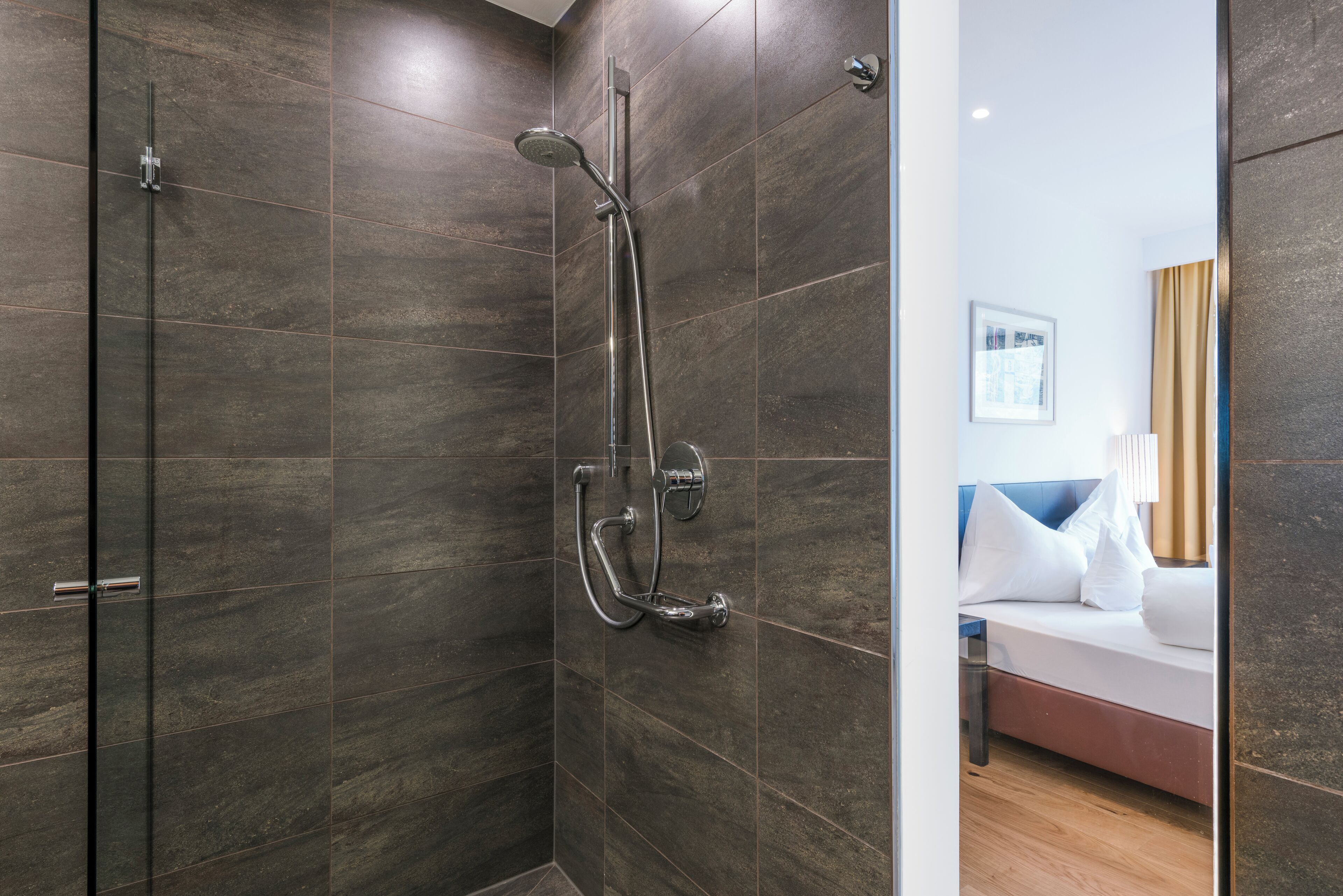 Une salle de bain moderne avec des carreaux de couleur sombre, une douche en verre et une chambre visible avec un lit et une table de chevet.