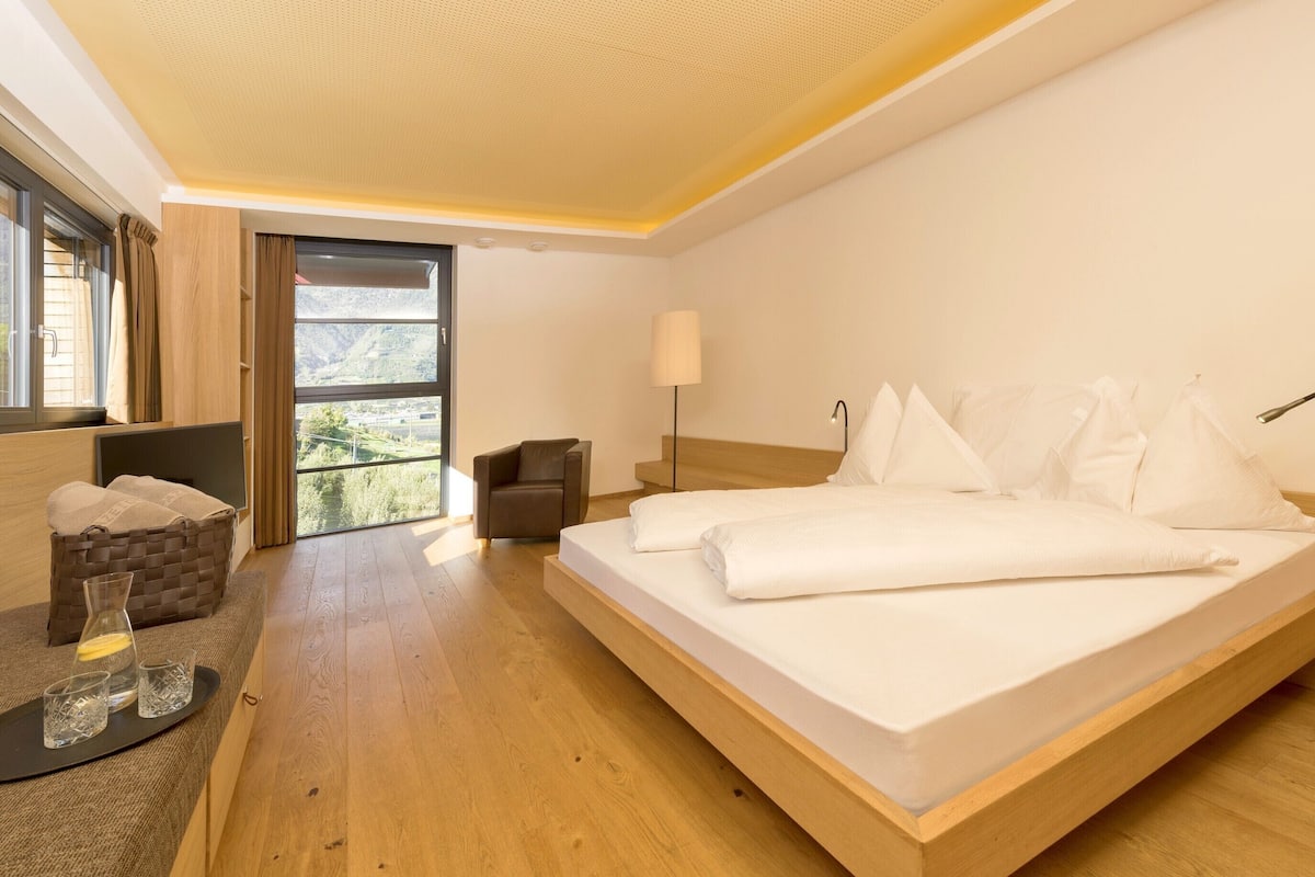 Una habitación de hotel moderna con una cama grande, un área de descanso con una silla, una mesita con vasos y una botella, y vistas a través de un ventanal amplio.