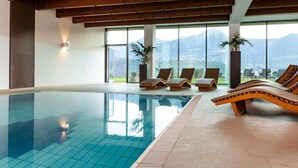 A large indoor swimming pool with clear blue water, surrounded by lounge chairs and large windows offering a mountain view.