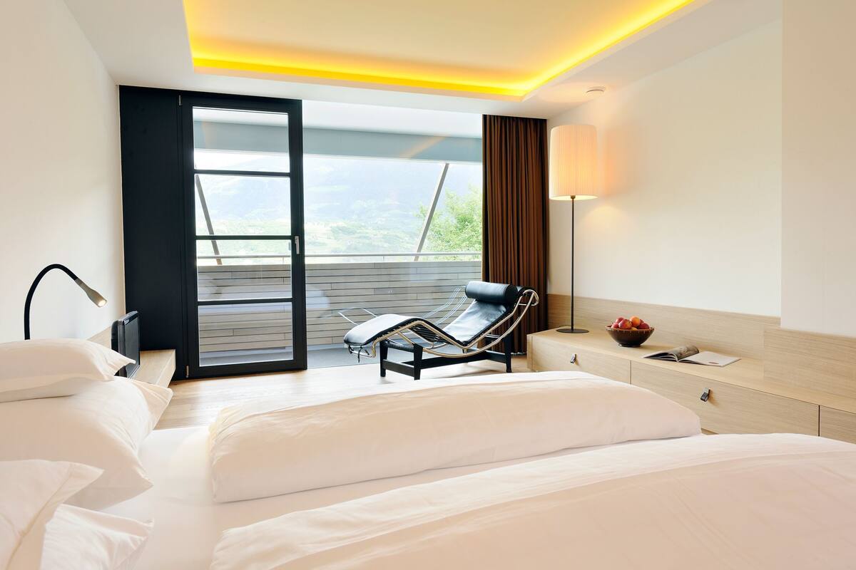 Un hotel moderno con cama, sillón reclinable, mesita de noche con lámpara y vistas al exterior.