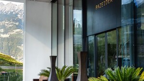 A modern building with a glass facade, a large outdoor seating area with a sofa and vases, and a mountainous view in the background.