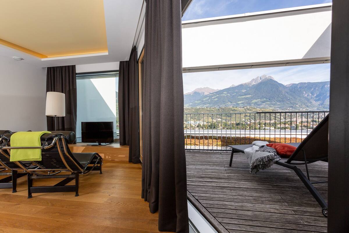Una habitación de hotel moderna con balcón, sofá, televisor y vistas a las montañas.