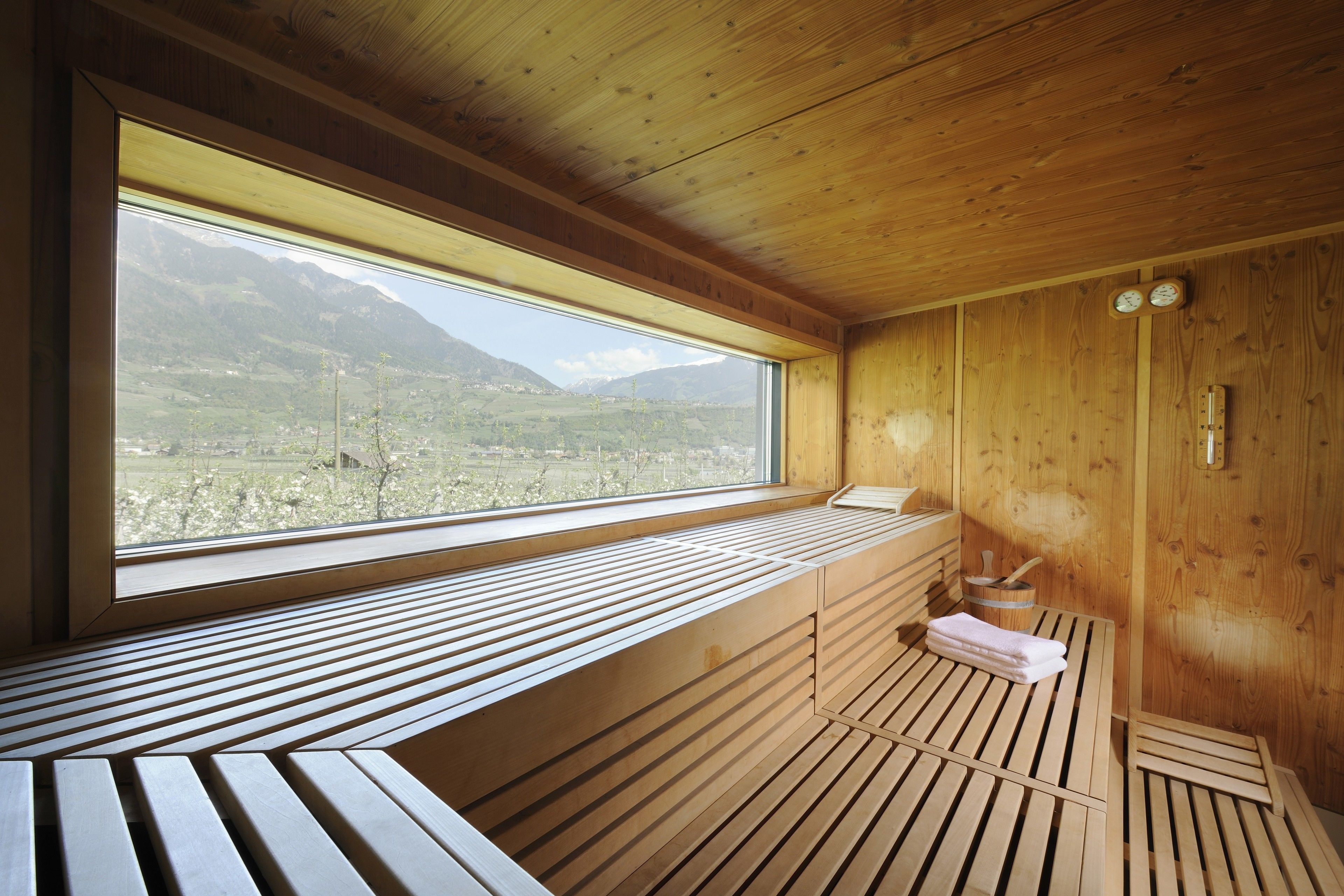 Una cabaña de madera con un ventanal grande que ofrece vistas a las montañas. En el interior, hay un banco de madera y una mesita con una toalla.