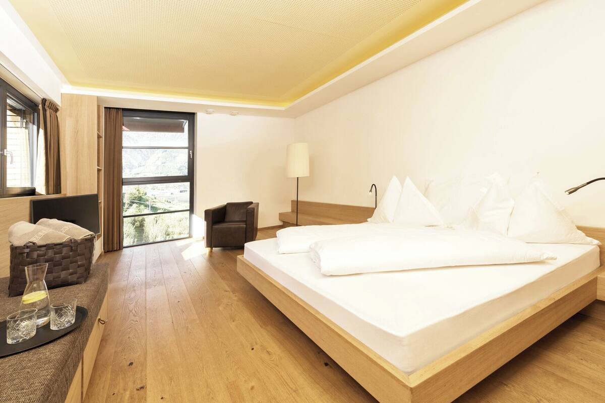 Una habitación de hotel moderna con una cama grande, una zona de estar con una silla, una mesita con vasos y vistas al exterior a través de un ventanal amplio.