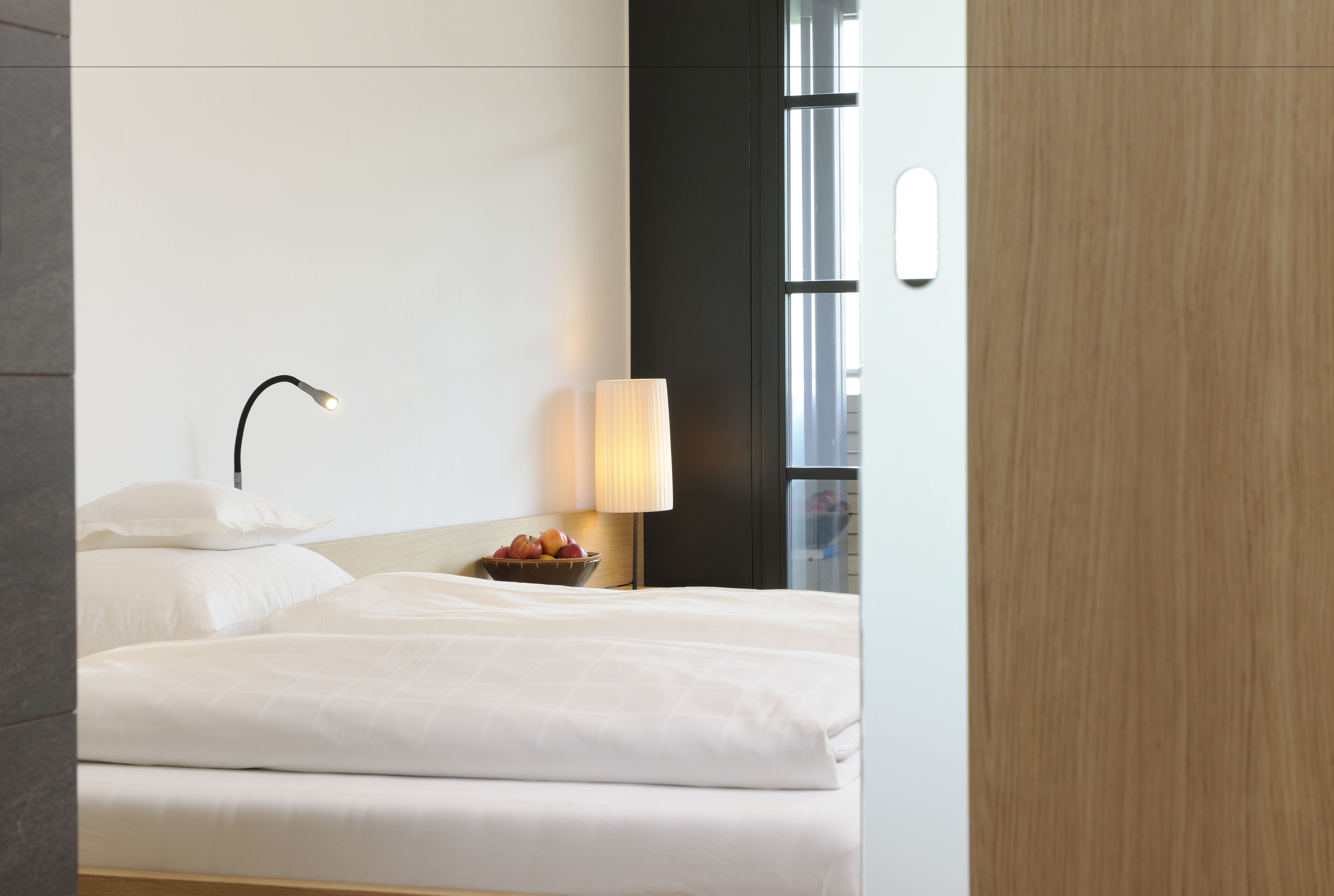 A hotel room with a neatly made bed, a bedside lamp, a bowl of fruit, and a window with blinds.