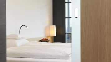 A hotel room with a neatly made bed, a bedside lamp, a bowl of fruit, and a window with blinds.