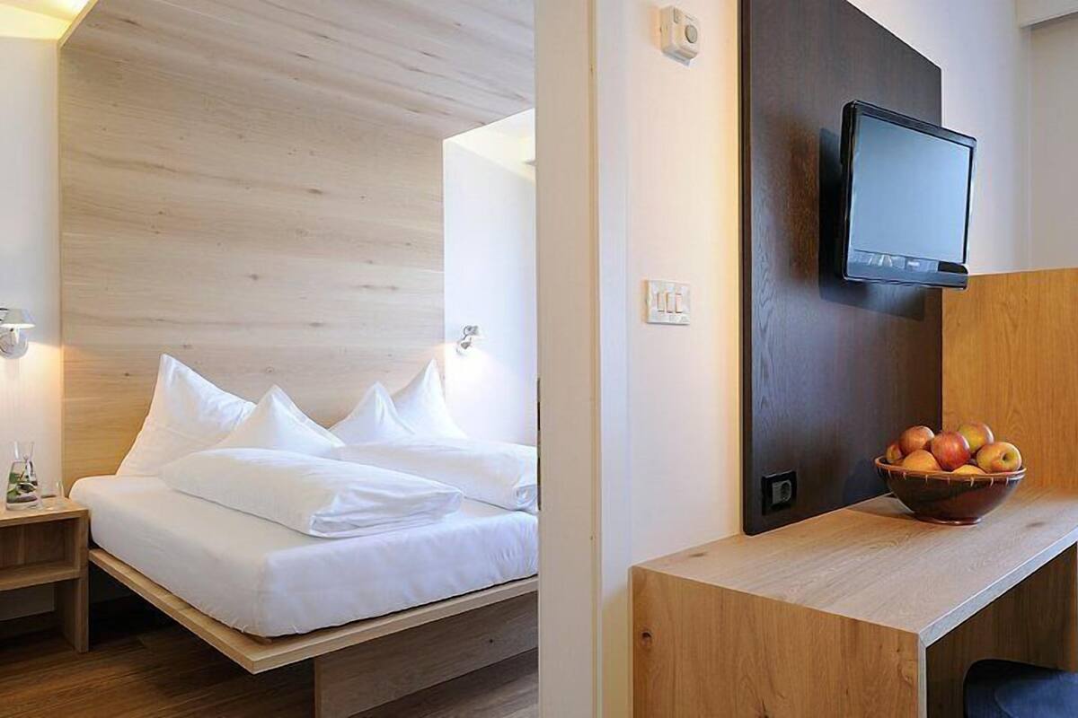 Habitación de hotel con cama, mesita de noche, lámpara, televisor montado en la pared y escritorio con frutero.