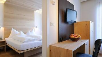 Une chambre d’hôtel comprenant un lit, une table de chevet, une lampe, un téléviseur fixé au mur et un bureau avec un panier de fruits.