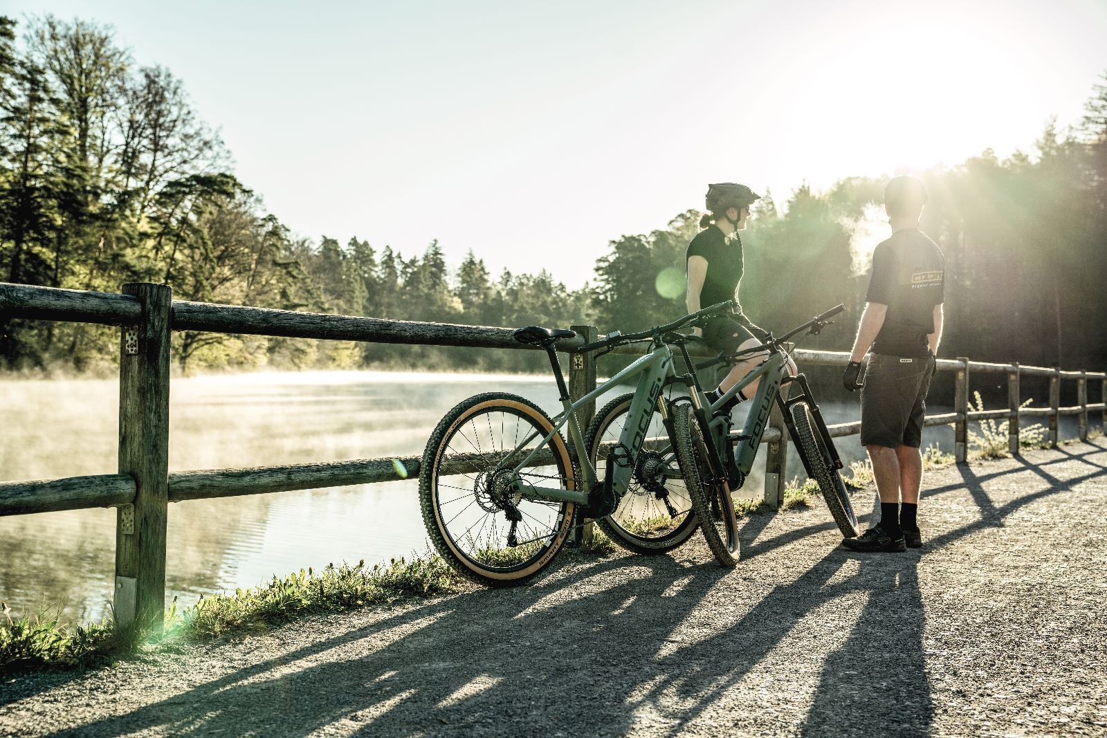 Eine Person steht neben einem Mountainbike auf einer Brücke, im Hintergrund ein See.