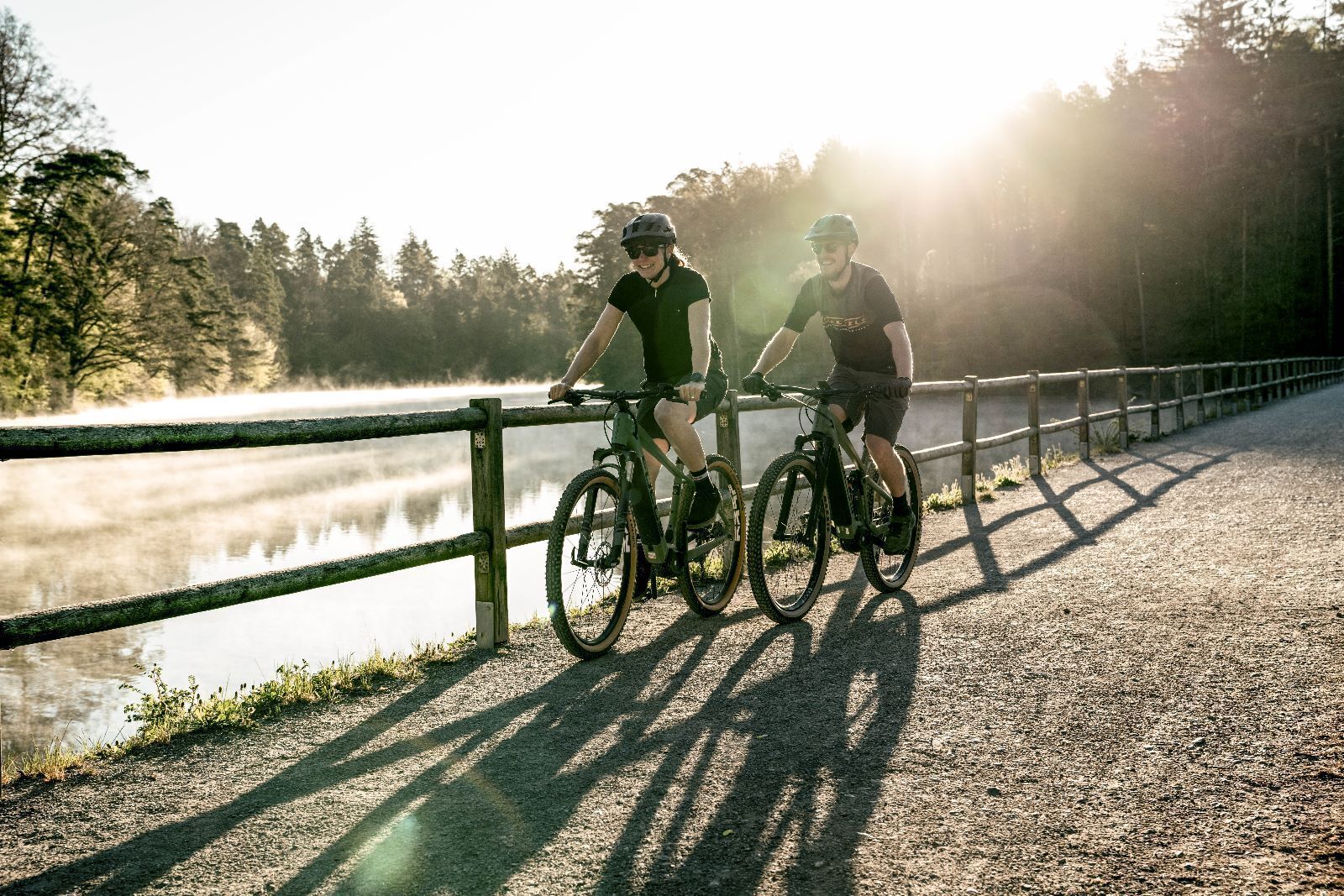 Two cyclists on a gravel path with a wooden fence and a body of water in the background.