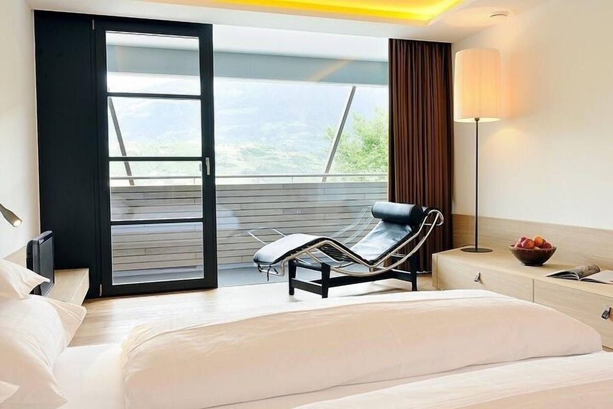 Una habitación de hotel moderna con una cama, un sillón, un escritorio y un ventanal con vistas a zonas verdes.