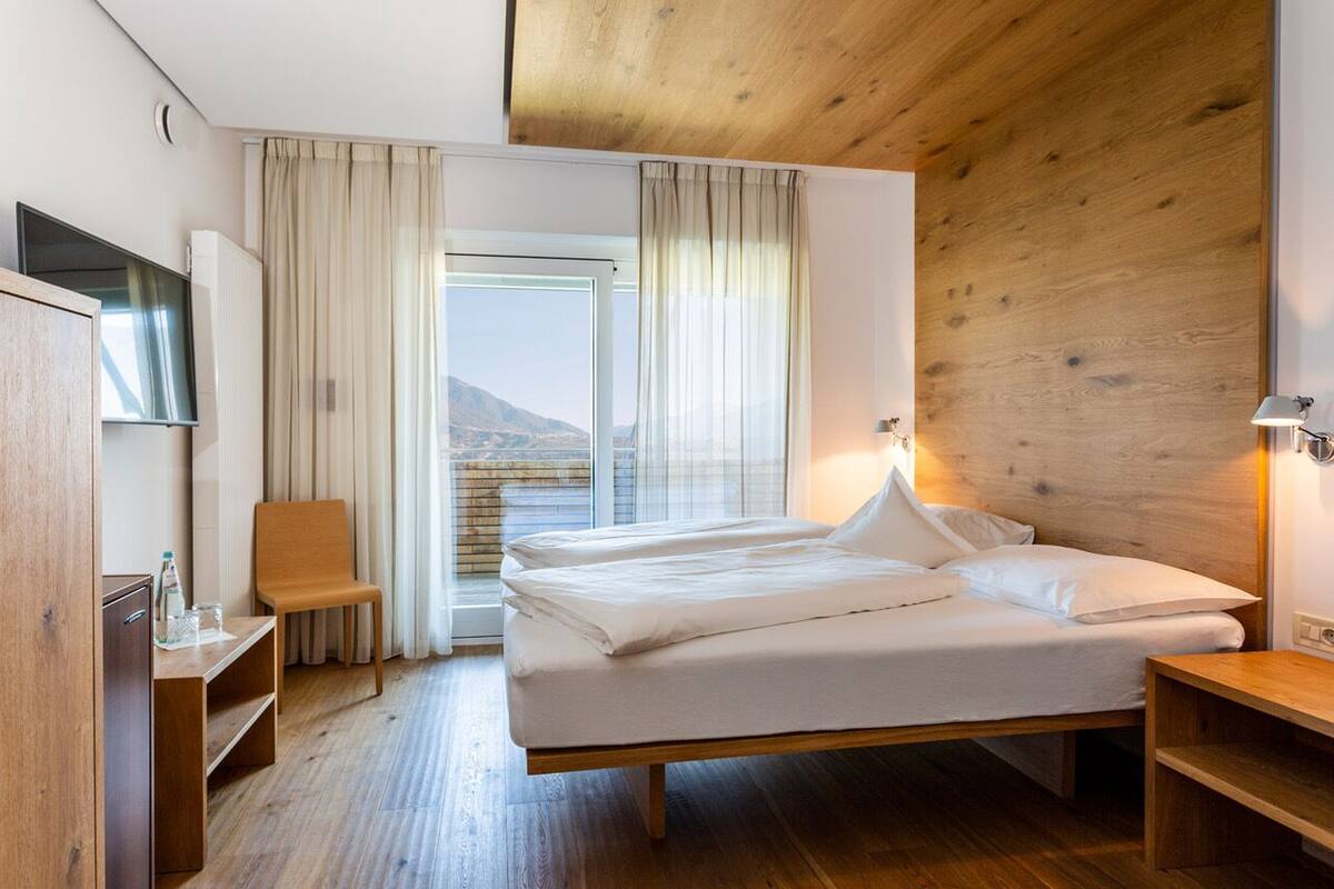 Habitación de hotel moderna con techo de madera, una cama grande con ropa blanca, televisión, escritorio y vistas a las montañas a través de la ventana.
