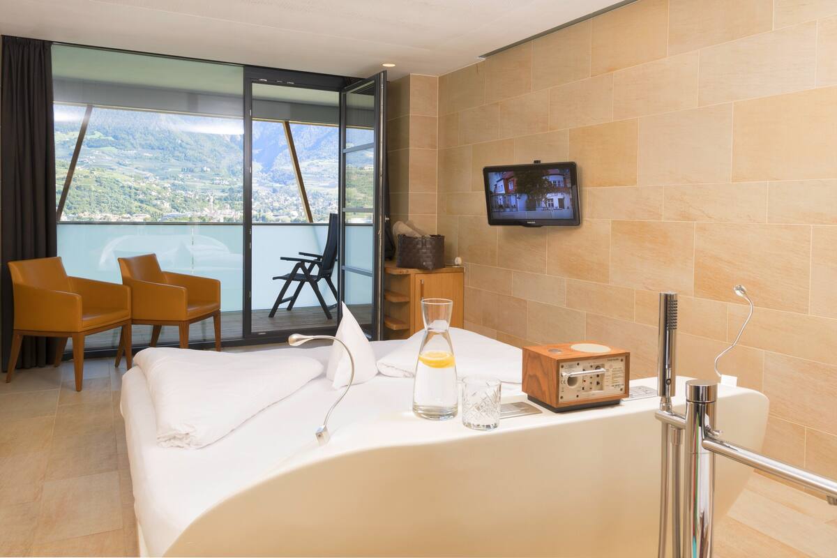 Habitación de hotel con una cama grande, un televisor en la pared, una silla, una mesita y vistas a las montañas a través de la ventana.