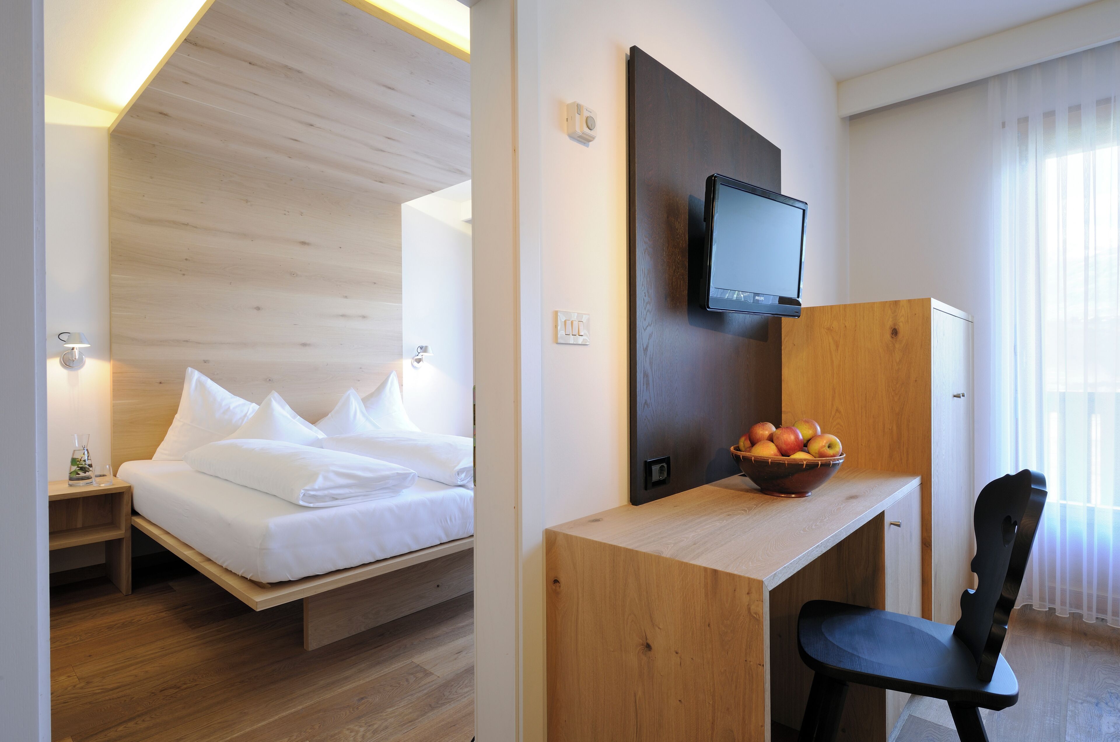 Habitación de hotel con una cama, un escritorio con una silla, un televisor y un frutero con fruta.