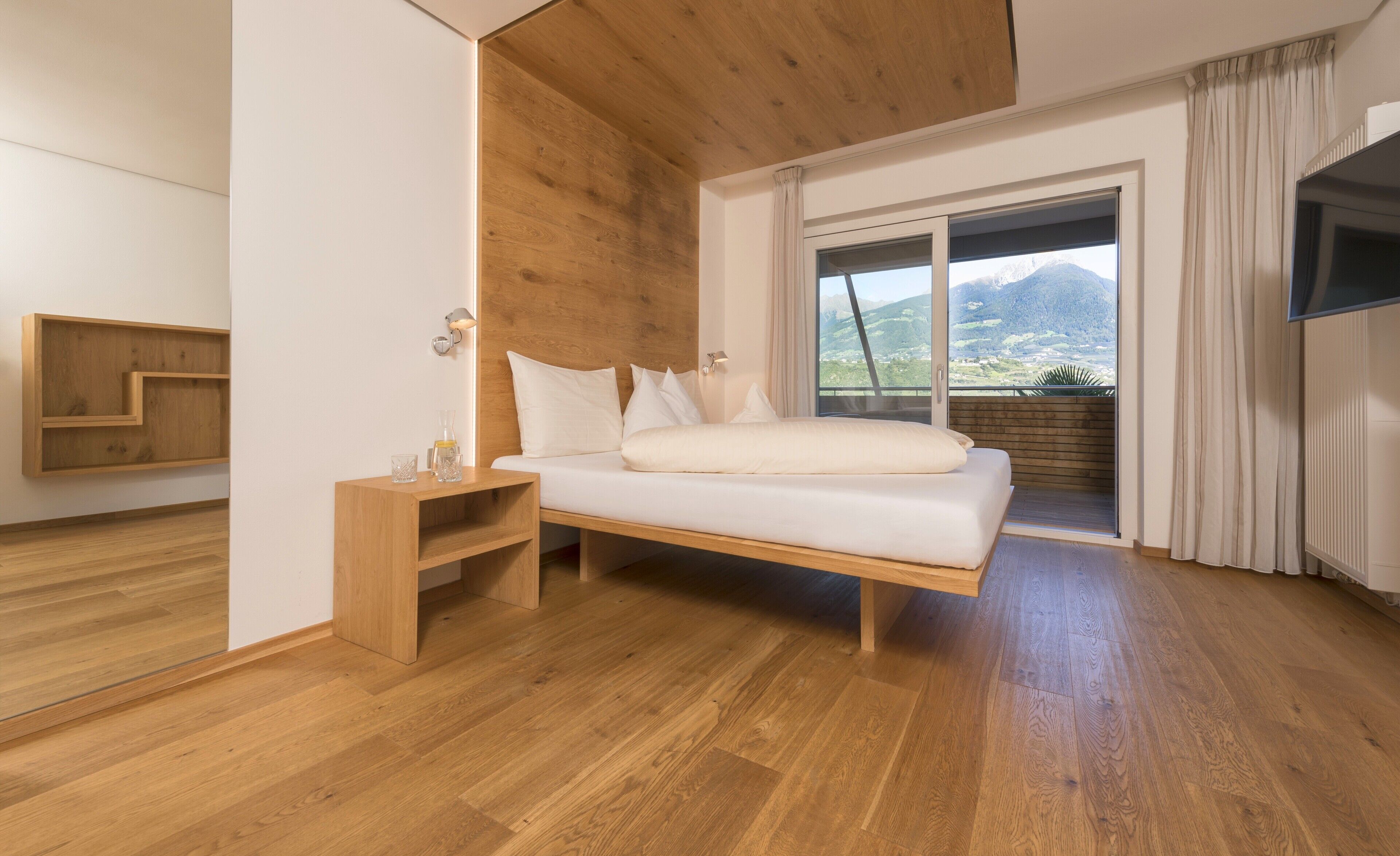 A modern bedroom with a wooden headboard, a built-in bench, a bed with white bedding, and a large window offering a mountain view.