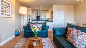 A living room with two sofas, a coffee table, and a vase of flowers. There is a kitchen in the background. - 2 Bed in Ilfracombe (oc-harbh) (Ilfracombe)