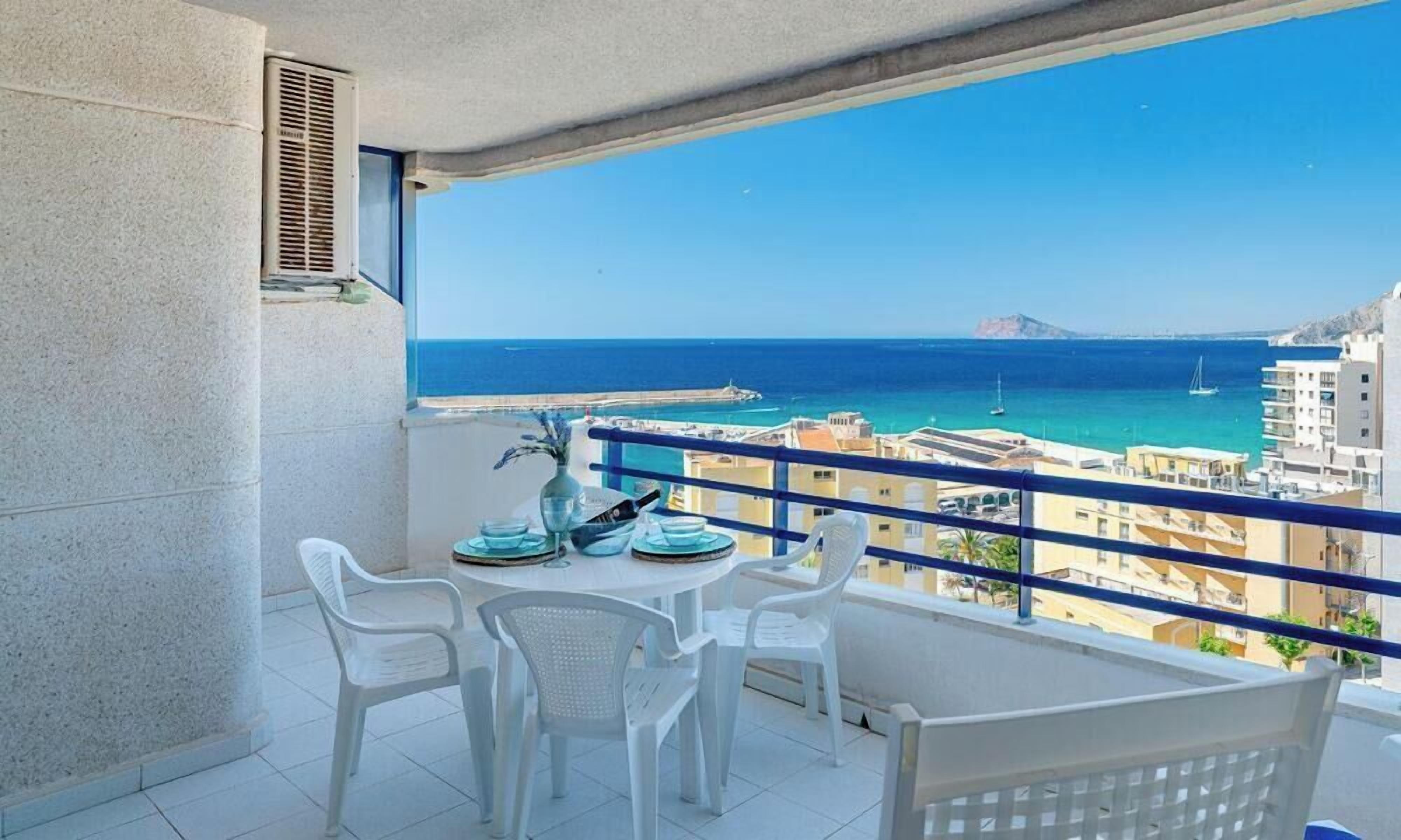 A balcony with a table and chairs, overlooking a coastal view with buildings and a distant island.