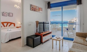 A hotel room with a bed, a TV, a small table, a sofa, a balcony with a view of the sea, and a window with curtains.