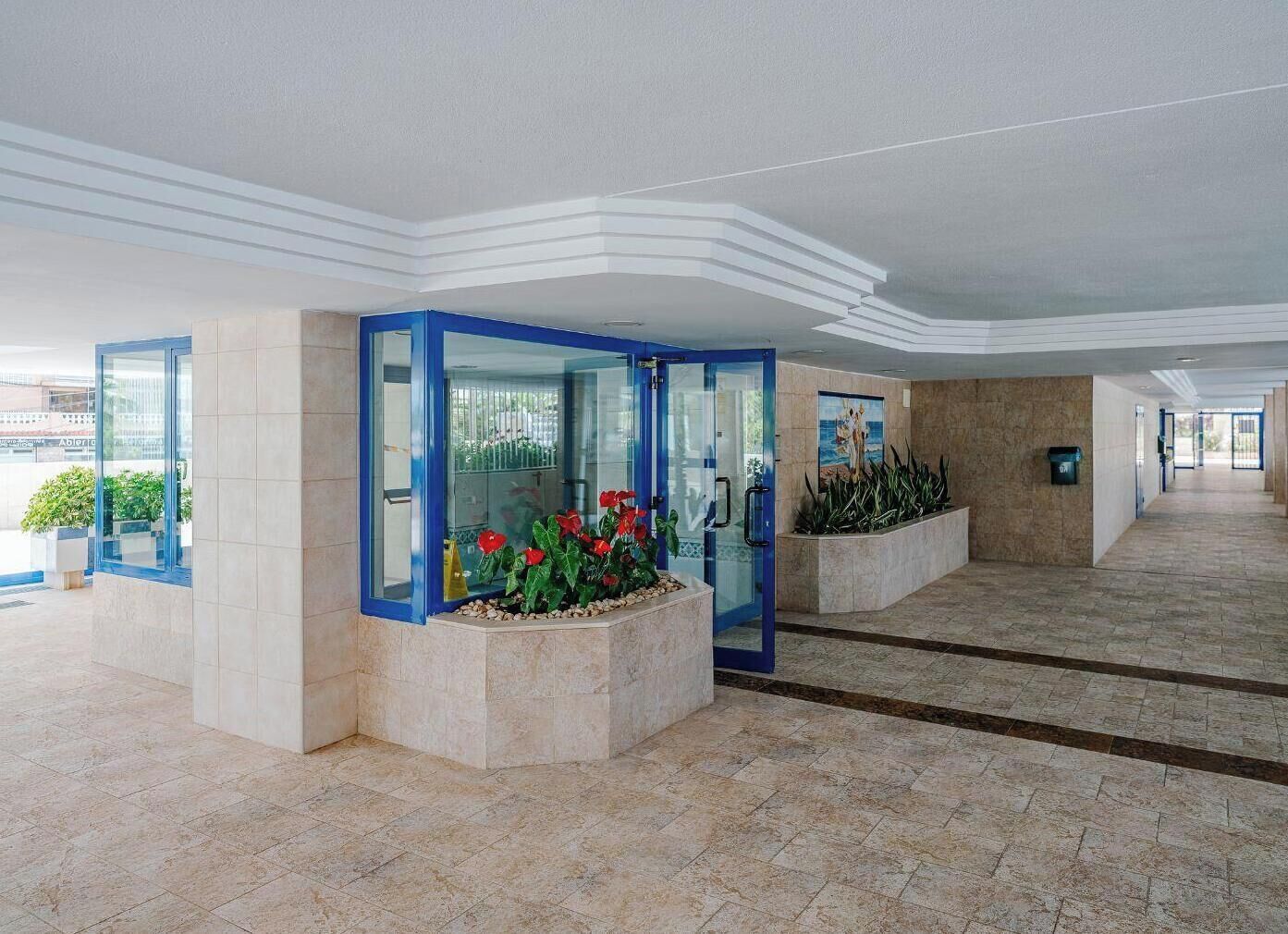 Un vestíbulo moderno con entrada por puerta de vidrio, piso de baldosas y un gran macetero con flores rojas.