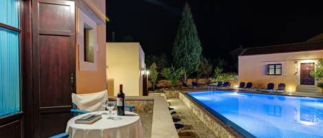 Un área junto a la piscina con mesa y sillas, una piscina y un edificio con puerta y ventanas.