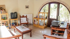 A cozy living room with a wooden chair, a small table, a bookshelf, a television, and a desk with a printer. - Heliopetra studios, small village close to beaches (Vamos)