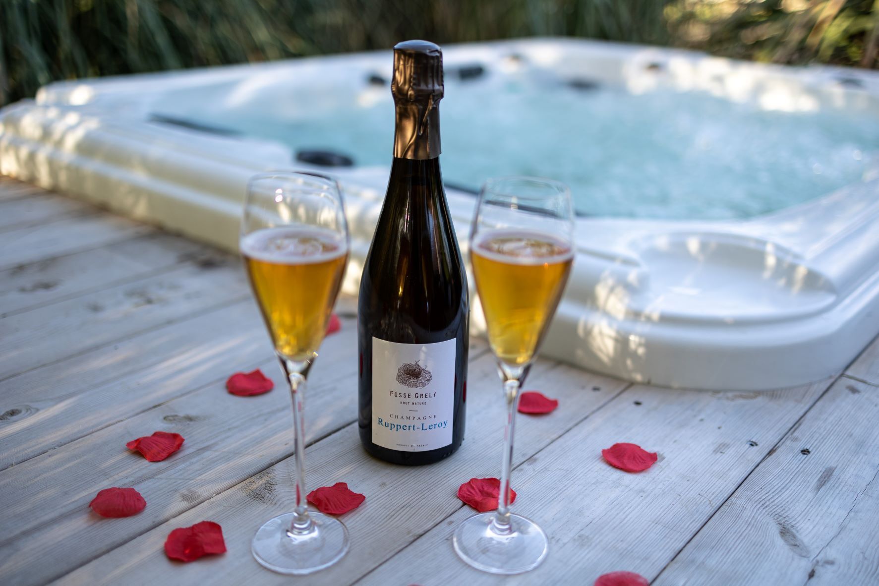 A bottle of Fosse Grely Brut Nature champagne, two glasses of champagne, and a hot tub on a wooden deck with rose petals.