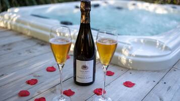 A bottle of Fosse Grely Brut Nature champagne, two glasses of champagne, and a hot tub on a wooden deck with rose petals.
