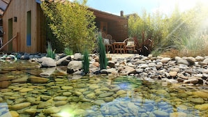 A wooden building with a tiled roof, a pond with clear water and rocks, and a seating area with chairs and tables. - Les Granges de Jules (Lafitte-Vigordanne)