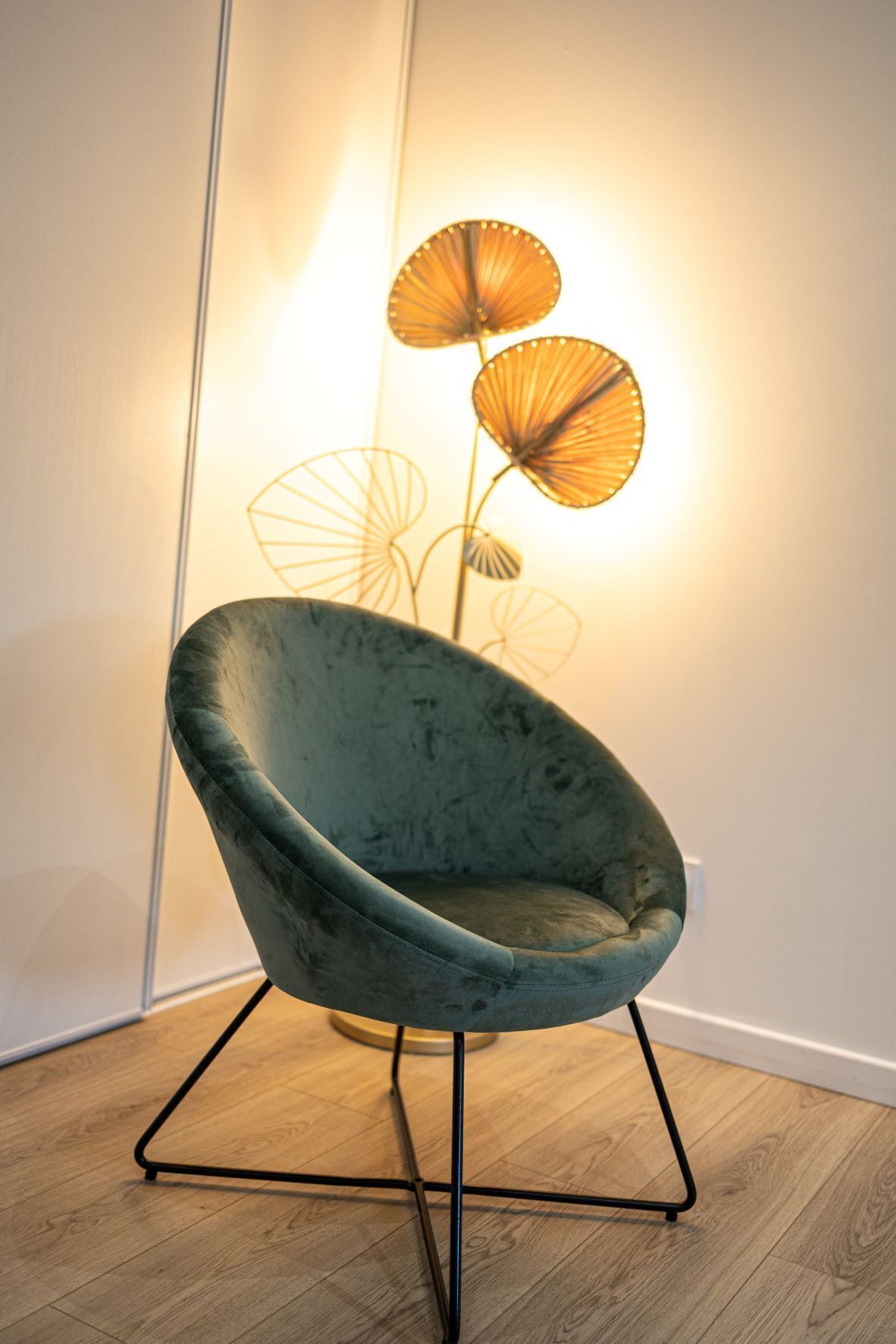 A teal velvet chair with a metal frame, positioned on a wooden floor against a white wall.