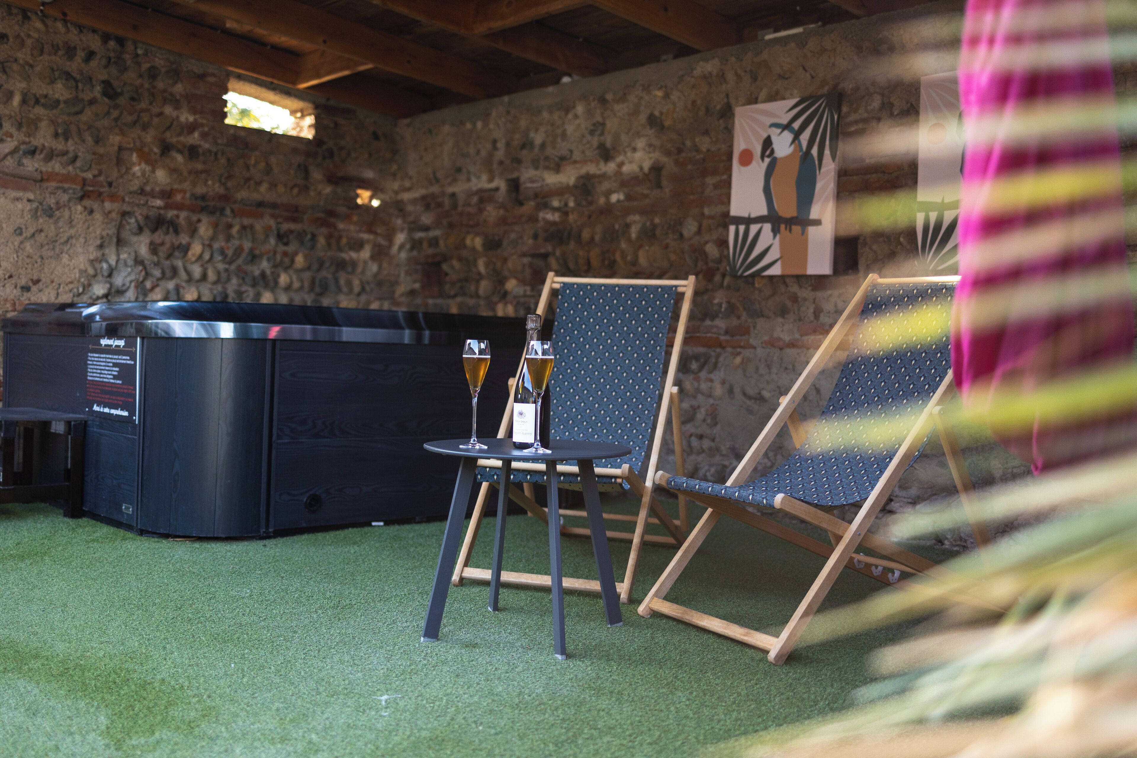 A stone-walled room with a hot tub, two folding chairs, and a small table with glasses.